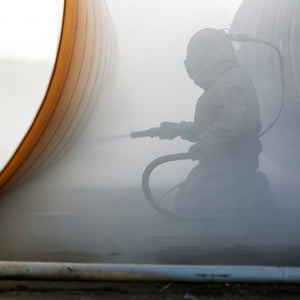 Person wearing a protective suit and helmet using a spray gun inside a large cylindrical structure.
